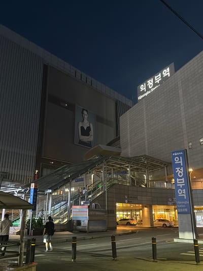 The image depicts the exterior of Uijeongbu Station in Uijeongbu-si, South Korea, at night. The station is a modern structure with a large building on the left displaying a billboard of a woman. Above the entrance to the station, a prominent sign reads "의정부역" in Korean and "Uijeongbu Station" in English. To the right, a blue vertical sign also displays "의정부역" and "Uijeongbu Station" along with some Chinese characters. Escalators and staircases lead up into the station, with people visible on them. Cars are parked in the illuminated area beneath the station entrance. The sky is dark, indicating it is nighttime, and streetlights illuminate the surrounding area. Several bollards with yellow reflective strips are positioned on the sidewalk.