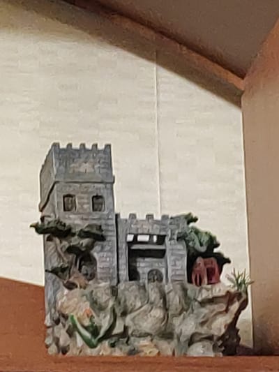 A diorama of a medieval castle is displayed on a shelf. The castle is made of grey stone and features multiple towers, battlements, and windows. It sits atop a rocky landscape with a small red structure that resembles a house or a smaller building on its right side. Green foliage is integrated into the diorama, appearing as trees and plants growing around the castle and on the rocky base. The background is a light-colored, textured wall. The shelf itself appears to be made of wood. The image does not provide any information to suggest its location in Brest, Belarus.