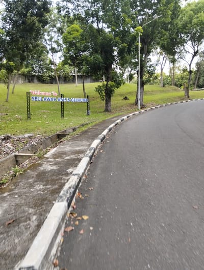 This media file depicts an outdoor scene, likely in Palohgadeng, Indonesia. The foreground is dominated by an asphalt road that curves to the right, bordered by a concrete curb with black and white markings. To the left of the road, there is a grassy area with some fallen leaves and a concrete drainage channel. Further back, a sign with the text "Welcome To SEULAWAll ATAS & GEURUTEE" is prominently displayed on a black metal frame. The sign has red lettering for "Welcome To" and blue lettering for "SEULAWAll ATAS & GEURUTEE". The background features lush green trees and foliage, with a concrete wall visible behind the grassy area. A lamppost stands among the trees to the right of the sign. The image appears to be taken during the daytime, with overcast or diffused lighting suggesting a cloudy sky. The overall impression is of a serene, possibly educational or recreational, environment. There are no people visible in the image, and the scene appears to be still. The quality of the image suggests it was taken with a handheld device, with some motion blur visible on the road and curb.