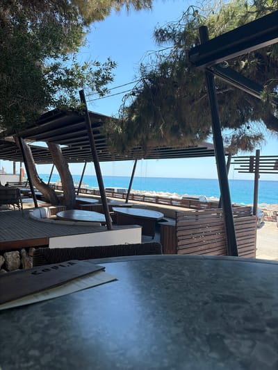 The image depicts an outdoor seating area of a beachfront establishment, likely a restaurant or cafe, in the Municipal Unit of Sfakiates, Greece. The foreground features a dark, textured tabletop with a menu book resting on it, displaying the word "KOPLA" embossed on its cover. Beyond the table, a wooden deck extends towards the sea, furnished with built-in seating and round tables. Two large tree trunks are integrated into the structure, supporting a dark-colored roof that provides shade. The middle ground showcases a picturesque view of the turquoise Ionian Sea under a clear blue sky, with a stretch of beach dotted with white umbrellas and lounge chairs. In the distance, a white vehicle is visible parked near the beach access. The overall scene conveys a relaxed atmosphere typical of a seaside resort.