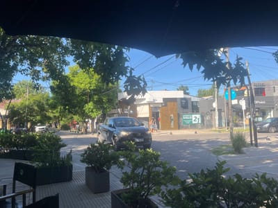 The image depicts a street intersection within City Bell, Argentina, observed from an outdoor patio area. The scene is illuminated by clear daylight under a bright blue sky. In the immediate foreground, dark outdoor planters containing green foliage and a tiled floor are partially visible, with a dark overhead structure (likely an awning or umbrella) obscuring the upper portion of the frame.
A dark grey pickup truck, possibly a Fiat Strada with its headlights illuminated, occupies the central midground, positioned as if traversing or turning through the intersection. Its Argentinian license plate, "DB 33X OC," is partially legible. To the left, a white car is visible further down the street. On the right side of the intersection, a dark sedan is visible near a building.
Pedestrians are present on the sidewalks: two individuals are observed on the left sidewalk near a building displaying a "UNIVERSIDAD SIGLO 21" sign. Additional pedestrian figures are discernible in the distance along the street. Dark, hemispherical bollards are installed along the curb lines at the intersection.
The surrounding environment consists of various commercial buildings. On the left, a building with a white facade and wooden accents prominently features the "UNIVERSIDAD SIGLO 21" signage. To its right, a dark grey building stands. Further right, another commercial building displays a "DPoint" sign. Numerous trees with dense green foliage line the streets, and utility poles with overhead wiring are visible against the sky, indicating a typical urban or suburban commercial thoroughfare.