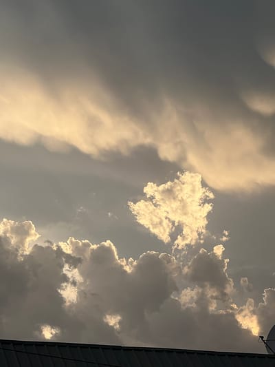 The image shows a cloudy sky above a dark, corrugated rooftop. The clouds are a mix of dark gray and bright white, with some areas illuminated by sunlight. The sky is mostly overcast, with streaks of lighter clouds against a darker background. There is no visible indication of the city of Balkh, Tajikistan in the image. The perspective is from ground level, looking upwards.