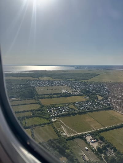 This is an aerial view captured from an airplane window, looking towards the city of Tårnby, Denmark. The scene is dominated by the vast expanse of the sky, with the sun prominently positioned in the upper left quadrant, casting bright rays and creating lens flare. Below, a body of water, likely the Øresund, stretches across the horizon, reflecting the sunlight.

The foreground is a patchwork of agricultural fields and a suburban settlement. The fields vary in color and texture, indicating different stages of cultivation. Interspersed within these fields are patches of woodland. The settlement is characterized by a dense collection of residential buildings with dark rooftops, arranged in a grid-like pattern. Roads and smaller pathways wind through the neighborhood. Further in the distance, industrial structures and more urban development are visible on the coastline.

In the lower right portion of the image, a small industrial or storage area is discernible, featuring several buildings, shipping containers, and a collection of vehicles, including what appear to be RVs or campers. Adjacent to this area, there are also signs of some discarded materials or a junkyard. No people are clearly visible in the image due to the aerial perspective and the scale. The overall impression is one of a coastal urban landscape with significant agricultural and natural elements surrounding it.
