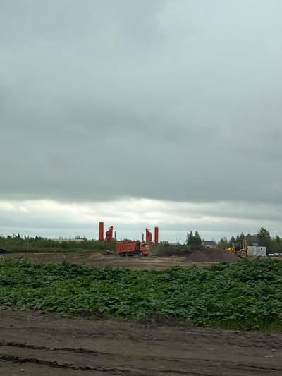 The image depicts a construction site near Myaglovo, Russia under an overcast sky. Heavy machinery including orange drilling equipment and a yellow bulldozer are present. The ground shows evidence of recent earthmoving activities. Vegetation grows along the edge of the construction area, bordering a dirt road or path. Two indistinct figures appear to be walking along the construction area. Buildings are visible in the background.