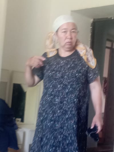 A woman wearing a dark, patterned dress and a white headscarf is standing in a room. She is holding a dark, folded object in her right hand and gesturing with her left hand towards her shoulder. A patterned scarf is draped around her neck and shoulders. The room appears to be indoors, with a light-colored wall behind the woman and a doorway on the right side of the frame. There is a reflection of a room or object on the left side of the frame, possibly a mirror or a window. The lighting suggests it is daytime. The location is identified as Mang'it, Uzbekistan.