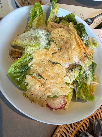 The image shows a Caesar salad on a white plate. The salad consists of lettuce, radishes, croutons, Parmesan cheese, and a crispy Parmesan cracker. The plate is sitting on a table, next to a menu and a woven placemat. The scene is inside a restaurant, possibly located in Oak Brook, United States of America.