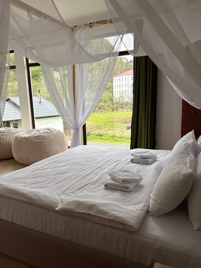 The image shows a bedroom scene, likely in a hotel or guest house, located in or near Khala, Georgia. Dominating the view is a bed with white linens, multiple pillows, and folded towels. A white, sheer canopy drapes around the bed, suggesting a romantic or luxurious setting. The window provides a view of the surrounding landscape, featuring a building with a light-colored facade and multiple windows, typical of residential or institutional architecture. Lush green vegetation fills the foreground of the outdoor scene. The window structure itself is modern, with dark frames. The lighting suggests it is daytime.
