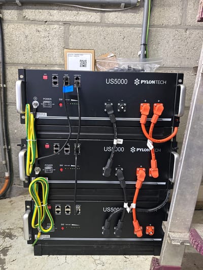 The image shows three stacked PYLONTECH US5000 battery units in what appears to be a utility room or storage area. The units are black with silver handles on the sides and have various ports and indicators on their front panels, including "POWER", "SW", "IN ALM", "CONTACT", "A/CAN", and "LINKPORT". Orange and black cables are connected to the battery units, with some labeled. There are also green and yellow ground wires connected to the units. A small cardboard box with a label containing "Article No: 3BW8W01-00084" and "Product: CABLES KIT" is visible on top of the uppermost battery unit. The background consists of a brick wall and some conduits. A metal ladder is partially visible on the right side of the image. The lighting suggests an indoor setting, likely with artificial light. There are no people visible in the image. The scene is clean and organized, indicating a functional installation.