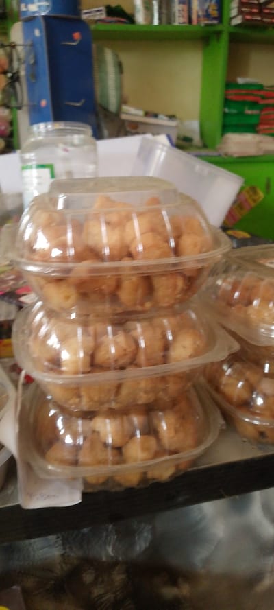 This image captures an indoor scene, likely a shop or market stall, located in Danbare, Nigeria. In the foreground, a stack of three clear, round plastic containers, each filled with numerous light brown, irregularly shaped baked goods or snacks, occupies the center. A fourth similar container is partially visible to the right. The containers are translucent, allowing the contents to be seen. Behind these containers, a clear glass jar with a white lid and a green label is visible. To the left, a dark blue and white machine or dispenser, possibly an office water cooler or a similar appliance, stands partially in view. Directly behind the items on the counter, a light yellow wall is visible, with green shelving units extending across the background. These shelves hold various indistinct objects, including what appears to be stacked fabric or packages in green and red, and other small items. A light-colored fan with a circular guard is partially visible on the left side of the background. The surface beneath the stacked containers appears dark and textured. No individuals are present in the frame.