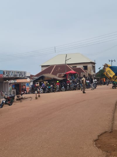 In Tangu, Nigeria, a bustling roadside scene unfolds with a group of motorcyclists gathered near shops. The street, paved with reddish-brown earth, is lined with various buildings, some appearing to be shops displaying goods. A red canopy is set up, offering shade over a display of items. Several motorcycles are parked in a row, with people standing around them. A man in a light-colored shirt and pants is walking away from the motorcycles. In the background, more buildings and power lines are visible against a hazy sky. A large blue and yellow banner, with text that includes "Dial," is prominently displayed on the right side of the image, partially obscuring some of the scene. A blue and red motorcycle is parked on the left side of the road. The overall impression is a lively, everyday moment in a Nigerian town.