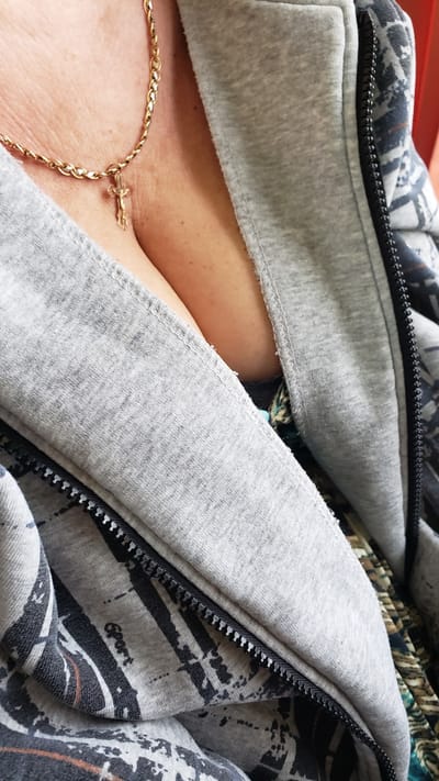 A close-up, eye-level shot focuses on the décolletage of a person wearing a gold chain with a crucifix pendant. The person is also wearing a grey hooded jacket with a zipper, revealing a glimpse of a patterned garment underneath. The background is out of focus and appears to be a red surface. The image contains no visible elements that would indicate a specific location within Podtureň, Slovakia.