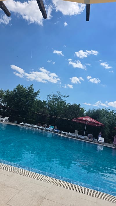 The image depicts an outdoor swimming pool area, likely located in Simeonovo, Bulgaria. The pool is filled with bright blue water and bordered by light-colored tiles and a slatted drainage system. Several white lounge chairs are arranged around the pool, with some occupied by people. In the background, lush green trees form a dense line, and a dark wooden pergola structure runs along the pool's edge. A large red umbrella with some visible text is situated near the right side of the frame. The sky above is a clear blue with scattered white clouds and a faint contrail from an aircraft. The overall scene suggests a sunny day at a resort or leisure facility.