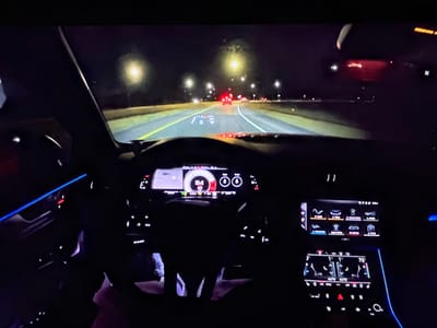 The image is a first-person view from inside a car at night, driving on a highway. The dashboard displays illuminated screens showing speed and navigation, with ambient blue lighting visible on the left side of the car's interior. Streetlights line the road, and other vehicles are visible in the distance, with their taillights illuminated. The sky is dark, indicating it is nighttime. There are no people clearly visible in the shot, except for a hint of someone's legs in the driver's seat. The car appears to be traveling at a speed of 84 mph, as indicated on the dashboard. The environment suggests a typical highway driving scene at night. No specific details about the city of Bezden, Bulgaria, can be inferred from the image.