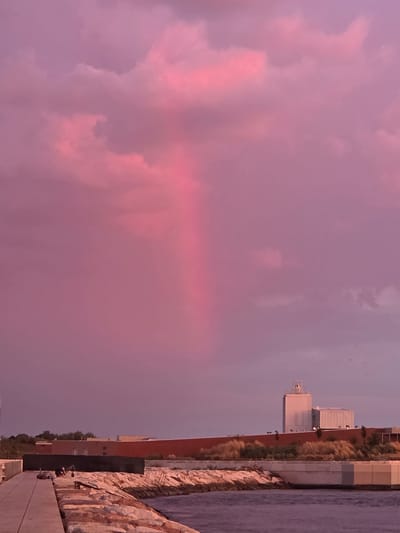 A vertical shot captures a vibrant pink sky over Chioggia, Italy. The sky is filled with soft, cotton-like clouds tinted with hues of magenta and fuchsia, with a distinct vertical shaft of light breaking through the clouds. In the distance, the silhouette of modern buildings, including a tall, rectangular tower, is visible against the horizon. A low, rust-colored wall runs along the foreground, separating the buildings from a rocky embankment. The embankment is made of rough, irregular stones and slopes down to the water. A walkway, possibly a pier or promenade, is visible on the left side of the image, made of light-colored rectangular planks. The water in the foreground is dark and calm, reflecting the colors of the sky. The overall scene evokes a sense of peacefulness and natural beauty, highlighted by the dramatic sunset.