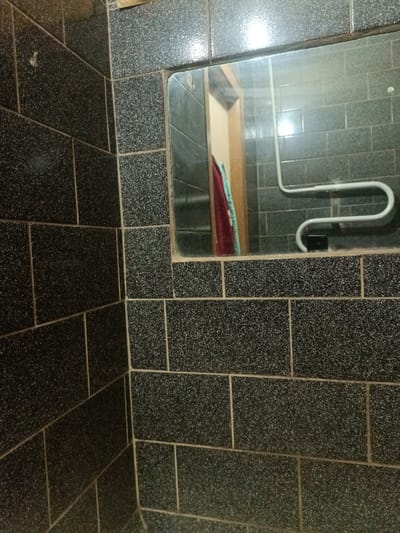 The image shows a corner of a tiled bathroom. The tiles are dark gray with a speckled pattern and appear to be roughly rectangular, laid in a brick pattern. The grout between the tiles is a lighter color. A mirror is mounted on one of the walls, reflecting another tiled wall and a towel rack. A red towel and a turquoise towel are hanging on the doorframe visible in the reflection. The overall impression is of a functional, perhaps dated, bathroom. The scene is indoors, likely a residential bathroom in Voskresenskiy, Russia, though no specific identifying landmarks are visible. There are no people present in the image.