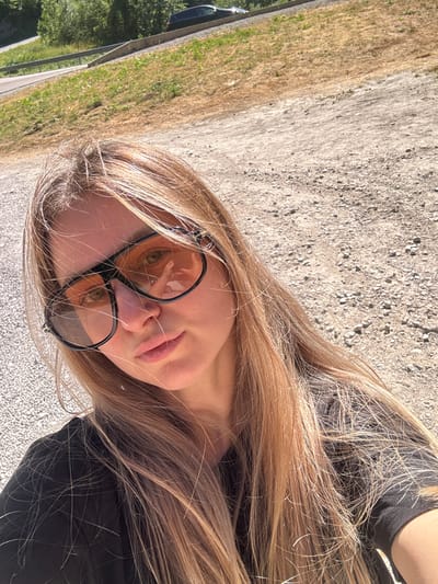 A woman with long blonde hair and light skin is taking a selfie outdoors. She is wearing large, dark-rimmed sunglasses with orange lenses and a black t-shirt. Her hair is windswept and partially obscures her face. The background consists of a gravelly surface, dry grass, and a road with a guardrail. In the distance, a dark-colored car is parked on the side of the road, and lush green trees are visible. The overall impression is a casual, sunny day. The location is identified as L'Essard, France.