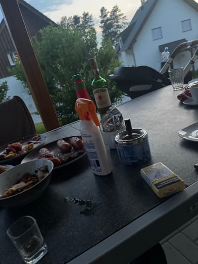 The image depicts an outdoor dining scene on a table, with food remnants and alcoholic beverages. In the background, a modern house is visible with some trees and a clear sky. On the table, there are plates of sausages and other food, a bottle of Jameson whiskey, a spray bottle, an ashtray with a cigarette, a pack of Camel cigarettes, and a glass. A baby stroller is also visible behind the table, along with a person standing near it. The location of Milná, Czechia, is not evident from the image.