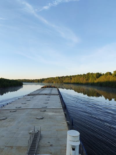 The image presents a serene long-shot perspective from the stern of a large vessel, likely a barge or a flat-deck cargo ship, as it glides along a calm river. The scene is dominated by a clear, expansive light blue sky with faint, wispy clouds overhead.
The foreground is occupied by the expansive, flat deck of the vessel, which appears to be constructed from grey, textured material, possibly concrete or steel. Numerous circular, recessed hatches or covers are evenly distributed across its surface. A metal staircase with an ornate, somewhat rusted railing descends from the current viewpoint towards a lower section of the deck. In the far end of the vessel, towards its bow, some stacked items, possibly cargo or equipment, appear in shades of yellow and orange. A white cylindrical structure, resembling a bollard, is visible at the bottom right corner of the frame. Blue railings line the sides of the deck, offering a view of the water below.
The river itself is tranquil, reflecting the clear sky and the dense green forests that line both banks. The vessel's movement is clearly indicated by the distinct ripples and a V-shaped wake trailing behind it, particularly prominent along the right side of the image. The trees appear lush and vibrant, suggesting healthy vegetation. In the distance, a subtle haze or mist seems to hover just above the water's surface, enhancing the peaceful atmosphere.
The lighting, with a soft golden glow touching the treetops on the right bank and the overall gentle illumination, strongly suggests the time of day is early morning. The weather is clear and calm, with no signs of precipitation or strong winds. The setting appears natural and remote. No discernible text is visible in the photograph. The location context provided is Srednii Rubezh, Russia.
