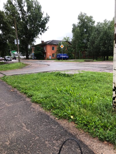 The image depicts a street scene in Safonovo, Russia, on an overcast day. In the foreground, a section of asphalt road curves to the left, bordered by a strip of overgrown grass. A white utility pole with a small sign is visible on the left side of the road. Beyond the road, an orange building with a dark roof and several windows stands amidst trees. In front of the building, a blue van with white lettering, likely a police vehicle, is parked. The road surface shows signs of wear and tear with patches of unevenness. The surrounding environment is characterized by lush green foliage and mature trees.