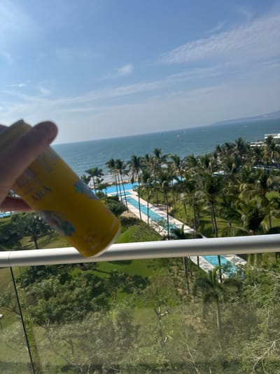 A yellow beverage can is held in a hand against a scenic backdrop. The view encompasses a blue ocean, a coastline with palm trees, and a resort-style swimming pool. Additional greenery and trees are visible in the foreground. A white, vertical railing element adds depth to the composition. The scene suggests a tropical vacation setting.