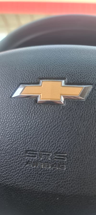 A close-up, vertical, medium shot captures the Chevrolet logo on a black leather steering wheel. The bowtie emblem, rendered in gold with a chrome outline, is centrally located. Below the emblem, the words "SRS AIRBAG" are embossed in the leather. The background is out of focus, showing a blurred pink and tan horizontal band, possibly a street sign or awning, and a curved gray surface, which could be part of the vehicle's interior or dashboard. The image is taken from within a vehicle, suggesting the perspective of a driver or passenger. The scene does not provide any specific information or landmarks that would allow for its localization to Muborak, Uzbekistan.