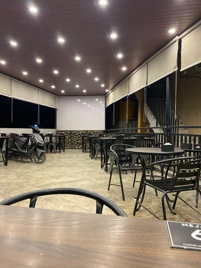 The image depicts an outdoor seating area, likely a café or restaurant, in Keudeublang, Indonesia. A row of black metal chairs and tables are arranged on a concrete floor, with some chairs stacked. A black motorcycle is parked in the seating area. Above, a brown ceiling with recessed lighting extends over the space. A sign with the word "MEJA" is visible on one of the tables.