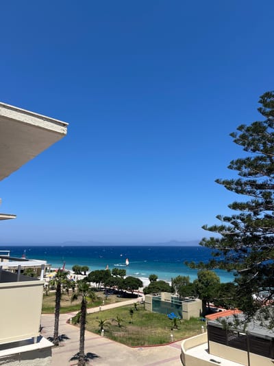 This image depicts a bright, sunny day at a coastal resort, likely in Rhodes, Greece. The scene is dominated by a vast expanse of clear blue sky, with the upper portion of the frame featuring the feathery fronds of pine trees, suggesting an outdoor setting.
Below the sky, the turquoise waters of the Aegean Sea stretch towards a hazy horizon. Along the coastline, there are signs of activity: several kitesurfers are visible on the water, their colorful sails dotting the waves. The beach appears sandy, with people scattered along its edge.
The foreground and right side of the image showcase a resort complex. Buildings with light-colored facades, red-tiled roofs, and balconies are visible. Green grass lawns are interspersed with pathways and palm trees, creating a well-maintained outdoor area. Some blue lounge chairs are set up on the grass, hinting at leisure and relaxation. The presence of the sea, kitesurfers, and resort buildings strongly suggests a vacation destination.
The time of day appears to be midday, given the bright sunlight and the clear, cloudless sky. The weather is clearly pleasant and warm, ideal for beach activities. There is no visible text in the image. The overall mood conveyed is one of tranquility and summer vacation enjoyment.