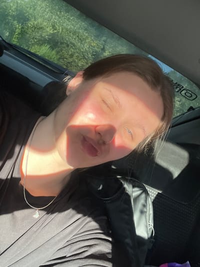 The image shows a close-up selfie of a young person inside a vehicle. The person is looking at the camera with their eyes partially closed and their lips pursed. Sunlight is streaming in from the side, casting a bright shadow across their face and illuminating their skin with a warm glow. They are wearing a black top and a silver necklace with a small pendant. The background shows the interior of a car, with seats and a bag visible. Outside the car window, green foliage can be seen, suggesting a natural, possibly rural setting. The strong sunlight indicates it is daytime, likely a sunny day. The overall mood appears relaxed and casual. On the car's interior panel, a logo with the letters "BR" and a circular symbol is visible. The setting is identified as Shuvakish, Russia.