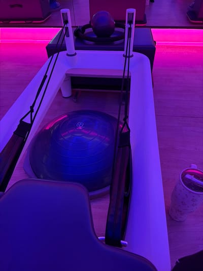 The image depicts a pilates reformer machine in a room with vibrant pink and blue lighting. The machine is predominantly white with a blue exercise ball placed within its frame. Black straps are attached to the reformer. Part of a chair is visible on the right side of the image. In the background, there's a water bottle with a patterned design. The specific location within the city of Enterprise, United States of America cannot be determined from the image.