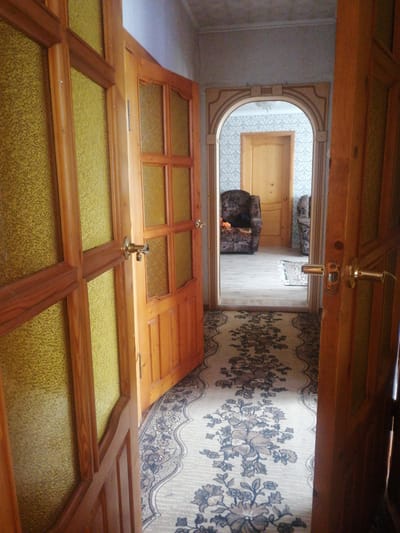 The image depicts an interior hallway of a residential dwelling. The scene is viewed from a low angle, looking down a carpeted hallway that extends into an arched doorway. The hallway is illuminated by overhead lighting, suggesting it is daytime or artificial lighting is used.
The primary subjects are architectural elements and furnishings. Two wooden doors with textured yellow glass panels are prominently featured. One door is open on the left, and another is open on the right. The hallway is adorned with a long, floral-patterned carpet runner. Beyond the arched doorway, another wooden door is visible, as are two upholstered armchairs and what appears to be a small rug on a light-colored floor. The wallpaper in the background has a subtle pattern.
No people are visible in the image, nor is any specific activity taking place. The notable details are the style of the doors, the patterned carpet, and the archway, all suggesting a traditional or older interior design. There is no visible text in the image. The overall impression is of a quiet, domestic interior space. The location context provided is Dubrovka, Russia.