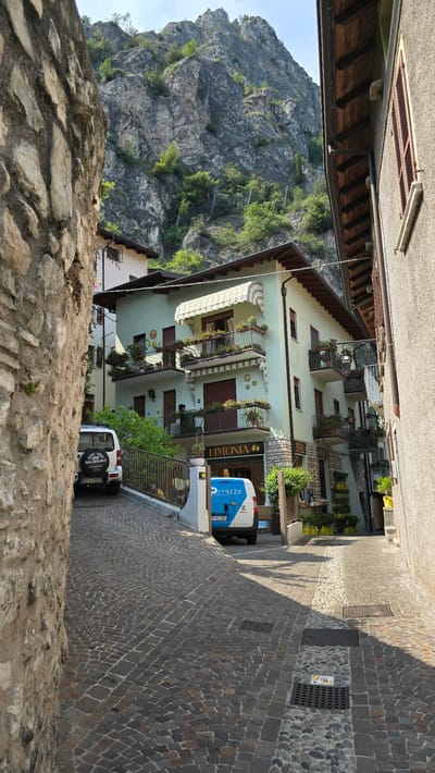 The image depicts a narrow cobblestone street in Limone sul Garda, Italy, leading uphill towards a steep, rocky mountainside. The architecture is characterized by traditional stone buildings, some with pastel-colored facades and balconies adorned with plants. A light green building with awnings and several balconies is prominent on the right side of the street, featuring a sign that reads "LIMONIA". Two vehicles are visible: a white Suzuki SUV parked on the left and a white and blue van with the text "purezza" on the right. The scene is bathed in sunlight, casting shadows across the cobblestones, and lush green vegetation can be seen growing on the mountainside and in planters on the buildings. The overall impression is of a picturesque Italian village nestled between mountains and water.