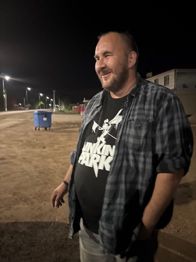The image shows a man standing outdoors at night. He is wearing a plaid shirt over a black t-shirt with white text. The text on the t-shirt appears to be "WATER PARK". The man is looking down and smiling, with one hand holding a cigarette. The background is dark, with some artificial lights visible in the distance, suggesting an urban or semi-urban setting. There are streetlights and a blue trash bin visible in the background. The ground appears to be dirt or gravel. The overall impression is a casual, late-night scene. The location context is provided as Kutlu-Bukash, Russia.