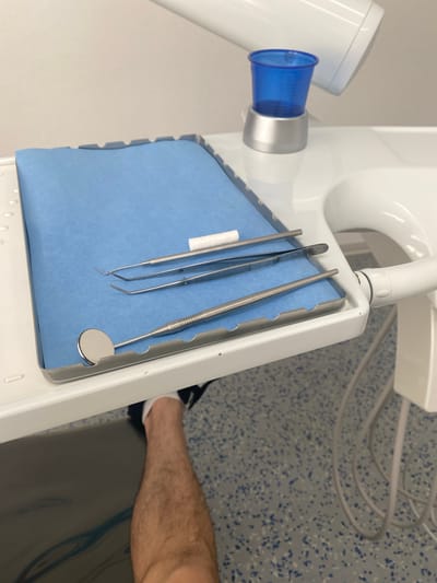 The image shows a close-up of a dentist's tray with instruments laid out on a blue paper. The tray holds several metal dental tools, including what appear to be tweezers, a probe, and a mirror. The instruments are arranged neatly on the blue paper. In the background, part of a dental chair or unit is visible, along with a blue suction cup and a white component. A person's leg, wearing white socks and dark shoes, is also partially visible in the lower right portion of the frame. The floor has a speckled pattern of blue and white. The overall setting is consistent with a dental examination room. There is no visible text in the image.