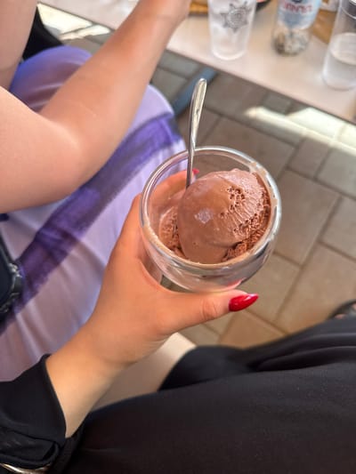 The image captures a close-up of a person holding a glass of chocolate ice cream. The hand, adorned with bright red nail polish, is gently cupping the glass. A spoon is partially submerged in the ice cream, suggesting it is being enjoyed. In the background, a person wearing black clothing is visible, and hints of a light-colored table and outdoor tiling suggest an outdoor setting. The lighting indicates it is daytime, likely with bright sunshine given the reflections and shadows. The overall mood appears relaxed and pleasant, as if someone is savoring a sweet treat on a nice day. No visible text is present on the ice cream or the glass.