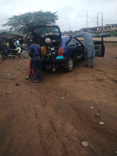 The image depicts an outdoor scene in Ajasse Ipo, Nigeria, characterized by a dirt road and a cloudy sky. A blue sedan with its trunk and passenger doors open is the central focus. Several individuals are present, including three young males, one seated in the open trunk and two standing near the rear of the car. One of the standing youths wears a blue t-shirt and dark trousers, while the other wears red trousers. Another young male, wearing a striped shirt and white cap, is seated inside the open trunk. A man in a light blue robe and trousers stands by the driver's side of the car, with his arms outstretched. In the background, a motorcycle with a rider is parked near a row of informal structures, possibly market stalls or shops. The ground is covered in reddish-brown dirt, with some tire tracks visible. Scattered leaves and debris are also present. The overall environment suggests a casual gathering or a temporary stop, possibly for loading or unloading goods.