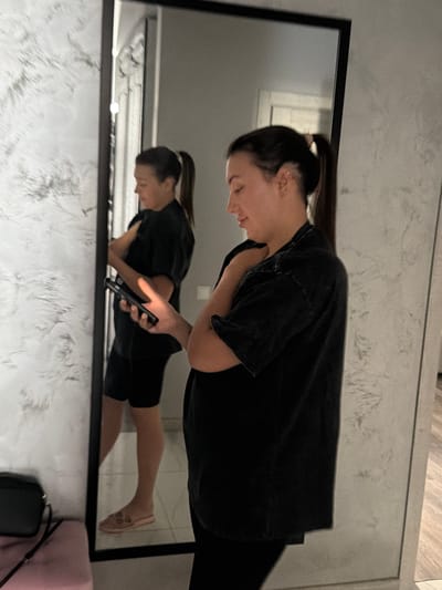 A young woman with her hair in a ponytail stands in front of a full-length mirror, looking at her phone. She is wearing a black t-shirt and black shorts. The mirror reflects her and a portion of the room behind her. The room appears to be a modern apartment with textured walls and light-colored flooring. A black handbag is visible on a pink surface to the left of the mirror. The scene suggests a personal moment captured in an urban domestic setting, possibly within the city of Novoaleksandrovka, Russia, given the contextual clue.