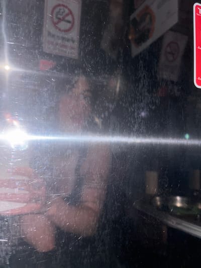 The image depicts an interior scene, observed through a heavily scratched, smudged, and reflective transparent surface. The location is identified as being within Rai Kao, Thailand, based on visible Thai script present on background signage.

A prominent horizontal glare streak from a strong light source bisects the lower-middle portion of the image, originating from the left. Numerous fine scratches and dirt particles are evident across the entire foreground surface.

In the background, visible through the obscured surface, are multiple signs. A "No Smoking" sign, circular with a red border and diagonal line over a cigarette icon, is clearly visible in the upper-left quadrant. A similar, fainter "No Smoking" sign appears in the upper-right. On the far right edge, a red rectangular sign with white Thai text and a partial logo (partially legible as "Yoon G") is present. Above the right-center, a rectangular sign, possibly a menu, displays dark text on a lighter background, with a faint image of a food item. The general background appears to be a dark-toned interior space, possibly a restaurant or bar, with a counter or shelf visible on the right, holding indistinct reflective objects.

Reflected in the foreground surface are at least two individuals. A person, appearing to be female with dark hair, is faintly reflected behind the central glare, with her face indistinct. Her right arm and hand are visible in reflection, holding a rectangular device, possibly a smartphone, and her fingernails appear long and painted. A portion of another person, likely the photographer, is reflected on the far left, appearing to interact with a red and white object.
