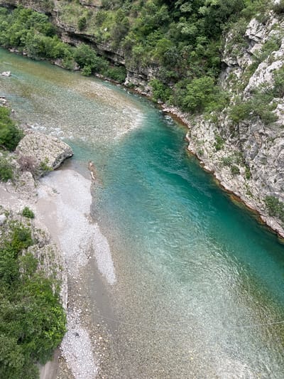 The image depicts a view from above of the Morača River canyon in Montenegro. The river is a clear turquoise color near the top of the frame, transitioning to a lighter shade as it flows downwards. Rocky cliffs densely covered with green vegetation border the river on both sides. The riverbed is visible in the shallower areas and consists of pebbles and rocks.