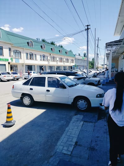 This is a photograph taken from a street-level perspective in Қарабұлақ, Kazakhstan. In the foreground, a white sedan is parked on the asphalt. To the left of the car, a yellow and black striped traffic cone is placed on the ground. Behind the sedan, a row of other vehicles, including white sedans, are parked. The background features a long, light-colored building with a green, sloped roof and multiple windows. Overhead, a network of electrical wires crisscrosses the sky. The sky is clear and blue with a few white clouds. A woman with long dark hair, wearing a white shirt and dark pants, is visible on the right side of the frame, walking away from the camera. Several other individuals can be seen further down the street, near the building entrance. The scene depicts a typical urban street with parked cars and commercial buildings on a sunny day.