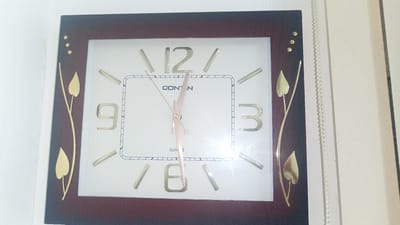 The image shows a square wall clock with a dark brown frame. The clock face is white with gold-colored numbers. The numbers 3, 6, 9, and 12 are clearly visible. The clock has three hands, hour, minute, and second, all of a pale pink color. The logo "CONT N" is present at the top of the clock face, and "QUARTZ" is visible at the bottom. Decorative gold-colored leaf motifs are attached to the left and right sides of the clock. The clock is mounted on a white wall. The provided context indicates the city of Maqat, Kazakhstan, however, there are no visual cues in the image to support or refute this location.