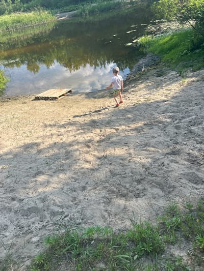 A child wearing a white t-shirt, green patterned shorts, a grey baseball cap, and red shoes stands on a sandy embankment next to a body of water. The child is facing away from the camera, towards the water. To the left of the child and closer to the water's edge is a wooden pallet. The water is calm and reflects the sky and surrounding trees. The embankment is composed of light-colored sand with scattered patches of green grass. The background consists of lush green foliage and trees. The scene appears to be a natural outdoor setting.