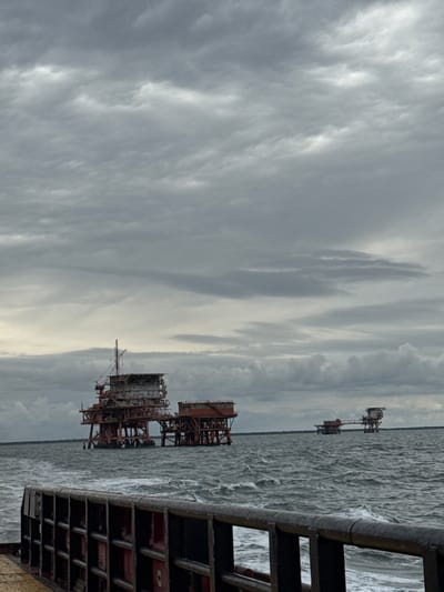 The image captures an industrial marine landscape under a heavily overcast sky. In the foreground, on the right side, a dark, weathered structure with horizontal and vertical beams creates a frame, suggesting the vantage point is from a pier or another platform.

The expansive body of water, likely the Adriatic Sea near Marina di Ravenna, Italy, is choppy with visible waves, indicating movement and perhaps wind. Several offshore platforms, possibly for gas extraction, are visible across the water. A large, reddish-brown platform with intricate scaffolding and structures dominates the mid-ground. Further in the distance, another similar but smaller platform can be seen, followed by a third, even smaller structure on the horizon, hinting at the vastness of the offshore operations.

The sky is filled with thick, textured grey clouds, allowing only muted light to filter through, suggesting an overcast day. The overall color palette is dominated by various shades of grey from the sky and water, contrasted with the rusty tones of the platforms. There are no people visible, and no specific activity or event is clearly taking place beyond the stationary presence of the offshore infrastructure. No discernible text is visible in the image.