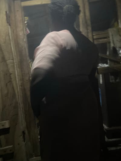 The image depicts a person viewed from behind, wearing a light pink shirt and dark clothing. The background suggests an indoor environment with wooden structures and possibly shelves. The lighting is dim, creating shadows and obscuring finer details. The scene appears to be within Omu Aran, Nigeria, though specific landmarks or contextual elements that confirm this location are not clearly visible in the provided image.