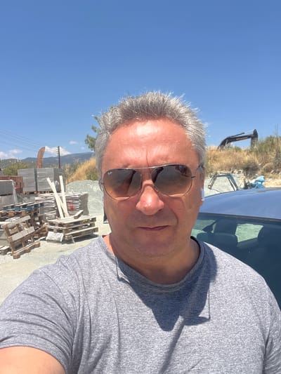 A man in his late 50s with graying hair and sunglasses is taking a selfie in front of a construction material yard in Laneia, Cyprus. He is wearing a gray t-shirt and white earbuds. In the background, there are stacks of bricks, wooden pallets, piles of gravel, and construction machinery. The sky is clear and blue, indicating a sunny day.