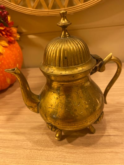 A close-up, eye-level, indoor shot focuses on a small, ornate brass teapot resting on a light brown wooden surface. The teapot is antique, with intricate etched patterns and a decorative lid topped with a finial. It has a curved handle and a slender spout. To the left of the teapot, a portion of an orange pumpkin with some red and orange decorative flowers is visible. The background shows a light-colored, textured wall with a woven wooden object partially obscuring the upper portion of the image. There are no people present in the scene. The lighting is warm and ambient, suggesting an indoor setting, possibly a home or shop. The scene does not provide any information about a specific city or country, therefore a location in Нарынка, Russia cannot be determined.