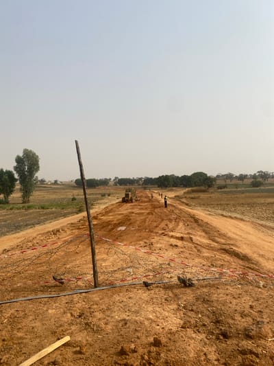 The image depicts an active road construction site in Wuciciri, Nigeria, under a hazy, light blue sky. A newly graded dirt road, reddish-brown in color, extends from the foreground into the far distance, flanked by flat, sparse fields and scattered trees.
In the immediate foreground, a rough, dark wooden post stands upright in the center-left. Coiled barbed wire lies on the ground around the base of the post, partially secured by red and white striped caution tape. A black pipe or cable runs horizontally across the lower foreground, beneath the barbed wire.
Midground details include a yellow construction vehicle, possibly a grader or bulldozer, positioned on the dirt road approximately midway down the visible stretch. Several individuals, at least four, are visible along the road beyond the vehicle, appearing to be workers; two in the mid-right wear high-visibility vests. The road surface shows tire tracks and disturbed earth.
The background features agricultural or fallow fields on both sides of the road, with clumps of green trees lining the horizon, indicating a rural or semi-urban environment. The overall impression is one of ongoing infrastructure development in an open, dry landscape.