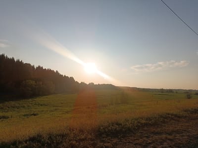 This is a landscape photograph taken in Alexandrovskoe, Russia, during sunset. The scene depicts a vast, open field bathed in the warm, golden light of the setting sun. A dense line of trees forms a dark silhouette along the horizon on the left side of the image, bordering a lush green meadow. The sky is a gradient of soft blues and oranges, with the sun itself a brilliant white orb directly in the center, casting long rays of light across the scene. A faint wispy cloud is visible in the upper right quadrant, and a thin power line stretches diagonally across the top of the frame. The foreground is composed of the uneven, brown earth of a dirt path or field edge, with dry grass and weeds. The overall impression is one of natural beauty and tranquility.