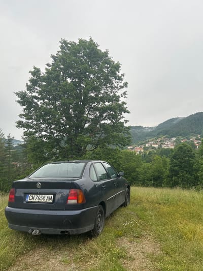 A dark blue Volkswagen Polo Classic sedan is parked on a grassy, slightly sloped area in the city of Ravnishta, Bulgaria. The car is viewed from the rear three-quarter angle, with its rear bumper, taillights, and license plate clearly visible. The license plate reads "CM 7658 AM". A large, mature deciduous tree with full green foliage stands to the left and behind the car, dominating the mid-ground. Beyond the tree, a village is nestled on a hillside, with houses featuring red-tiled roofs. The background consists of rolling, tree-covered hills under a cloudy, overcast sky, with some mist or fog present in the distance. The overall environment suggests a rural or semi-rural setting, possibly during or after a period of rain, as indicated by the slightly wet appearance of the car and the ground. The foreground is composed of dry grass, pebbles, and some small wildflowers. No people are present in the image.