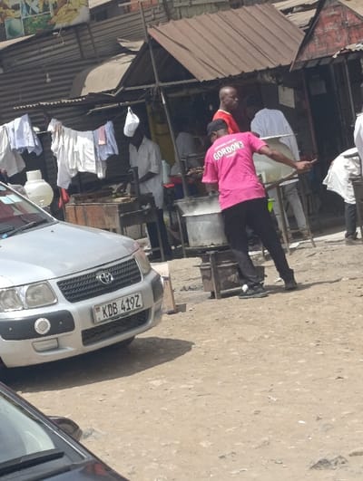 An outdoor scene in Matungulu west ward, Kenya, depicts a street food or market area under bright sunlight. The ground is unpaved, consisting of dirt and gravel.
In the foreground, the front bumper and grille of a silver Toyota Probox van, license plate KDB 419Z, are visible on the left, partially obscuring the front of another dark-colored vehicle.
In the midground, multiple individuals are present around makeshift market stalls. A male subject, wearing a bright pink t-shirt with "GORDON'S" printed in white on the back, a black baseball cap, and black trousers, stands facing away from the camera. He is actively stirring the contents of a large, metallic, cylindrical pot or vat, which rests on a metal stand, possibly a stove. To his immediate left, another male subject, bald and wearing a red t-shirt, stands observing the activity. Further left, a third male subject, dressed in a white collared shirt and possibly a white cap (hanging on a hook or his head), attends to a rectangular cooking station or grill. Behind him, white garments are hanging on a clothesline. Other individuals in light-colored clothing are visible within the background stalls.
The background features several informal structures with corrugated metal roofs, some reddish-brown, others grey. These structures appear to be stalls or small shops. Signage with imagery is partially visible in the upper left background. Shadows indicate direct overhead sunlight.