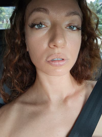 A close-up photograph features a person with fair skin, auburn curly hair, and light-colored eyes with prominent eyelashes. The person's facial expression is neutral, and they appear to be in an interior space, possibly a car, indicated by the presence of a seatbelt strap diagonally across their shoulder. There is no specific evidence to suggest the photograph was taken in Minh Tân Commune, Vietnam.